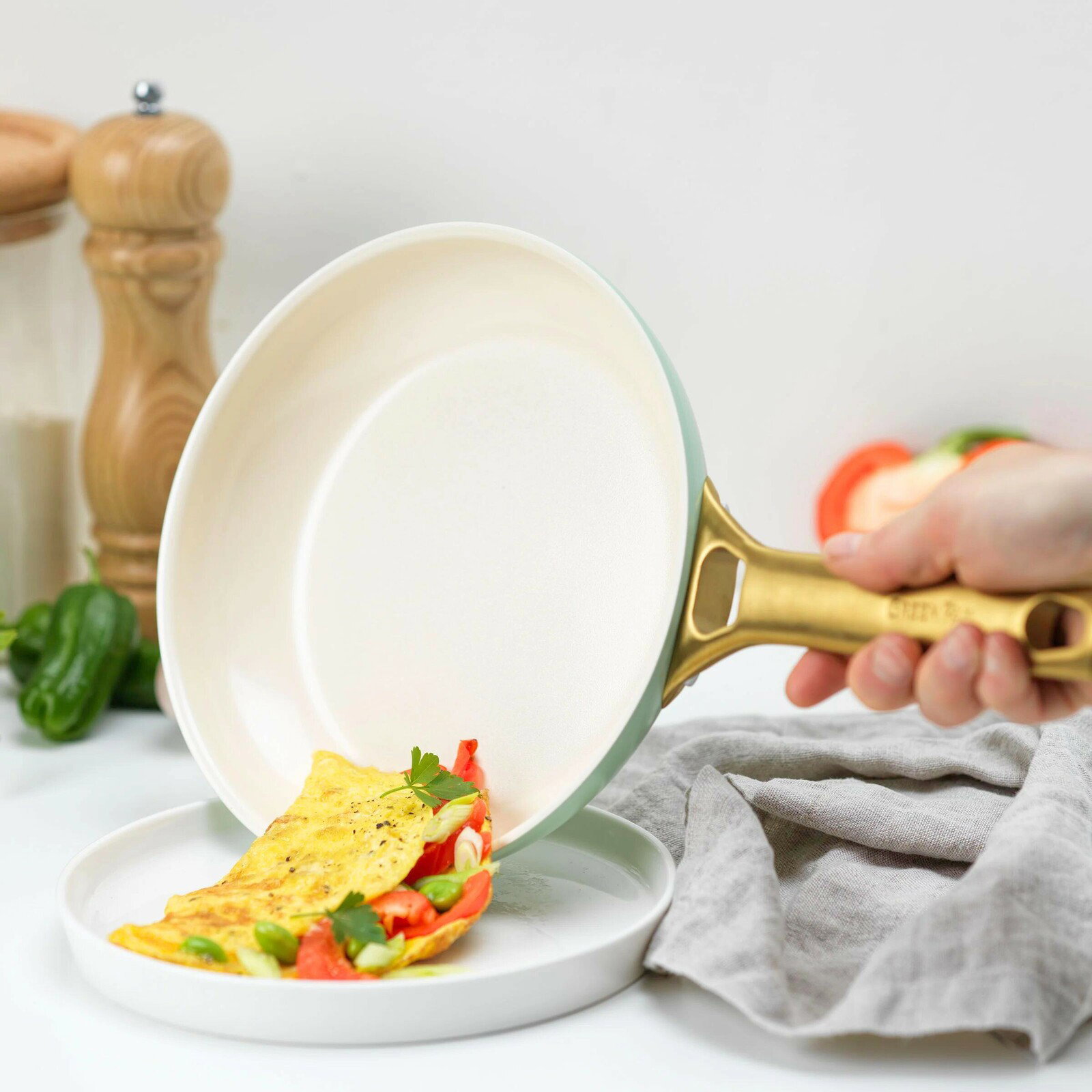 Nahaufnahme einer Bratpfanne mit goldfarbenem Griff, aus der ein Omelett auf einen Teller gleitet. Im Hintergrund sind Gewürze und Gemüse zu sehen.