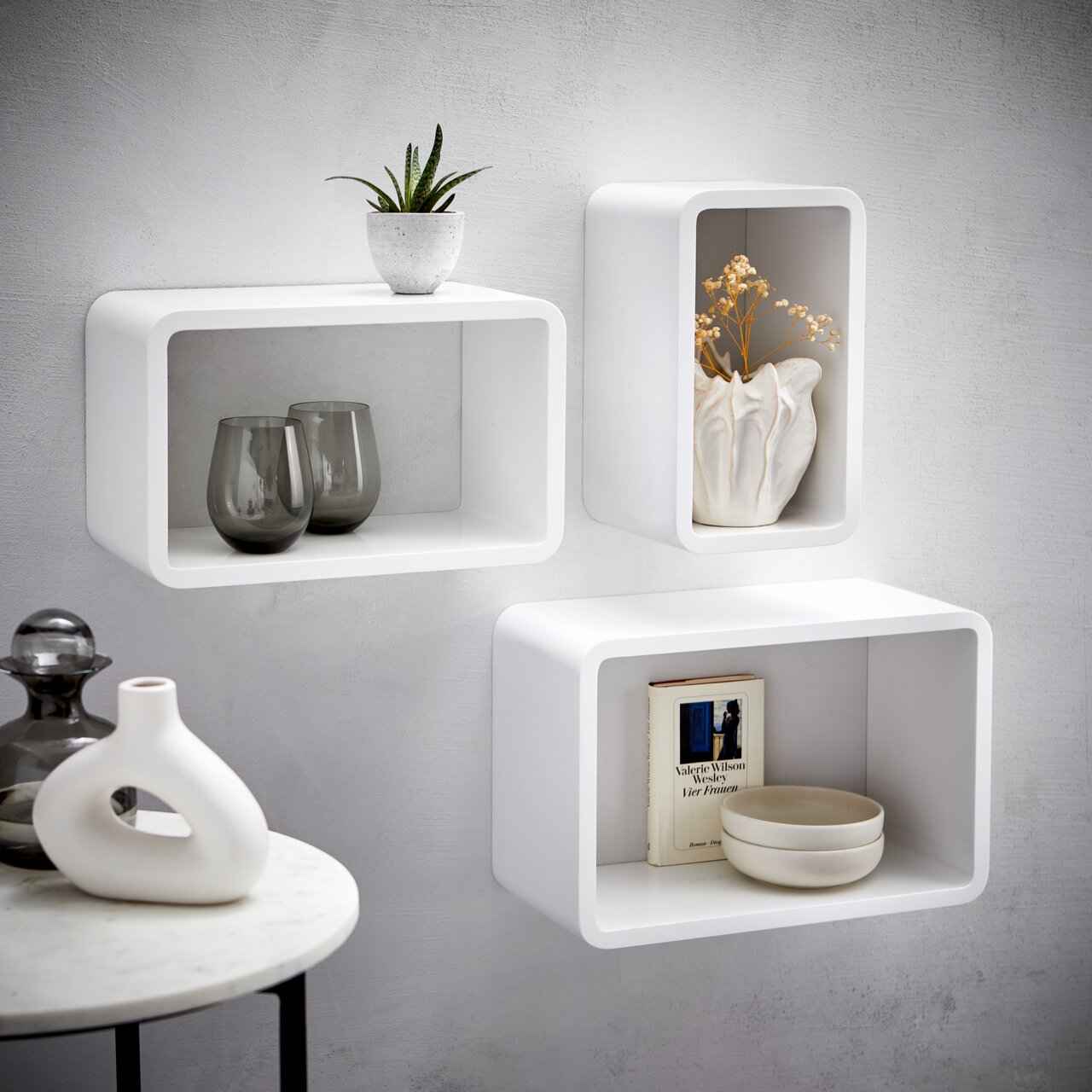 Wandregal-3er-Set in Weiß, bestehend aus drei rechteckigen Regalen, dekoriert mit Pflanzen, Vasen und Büchern, an einer grauen Wand montiert, seitliche Perspektive.