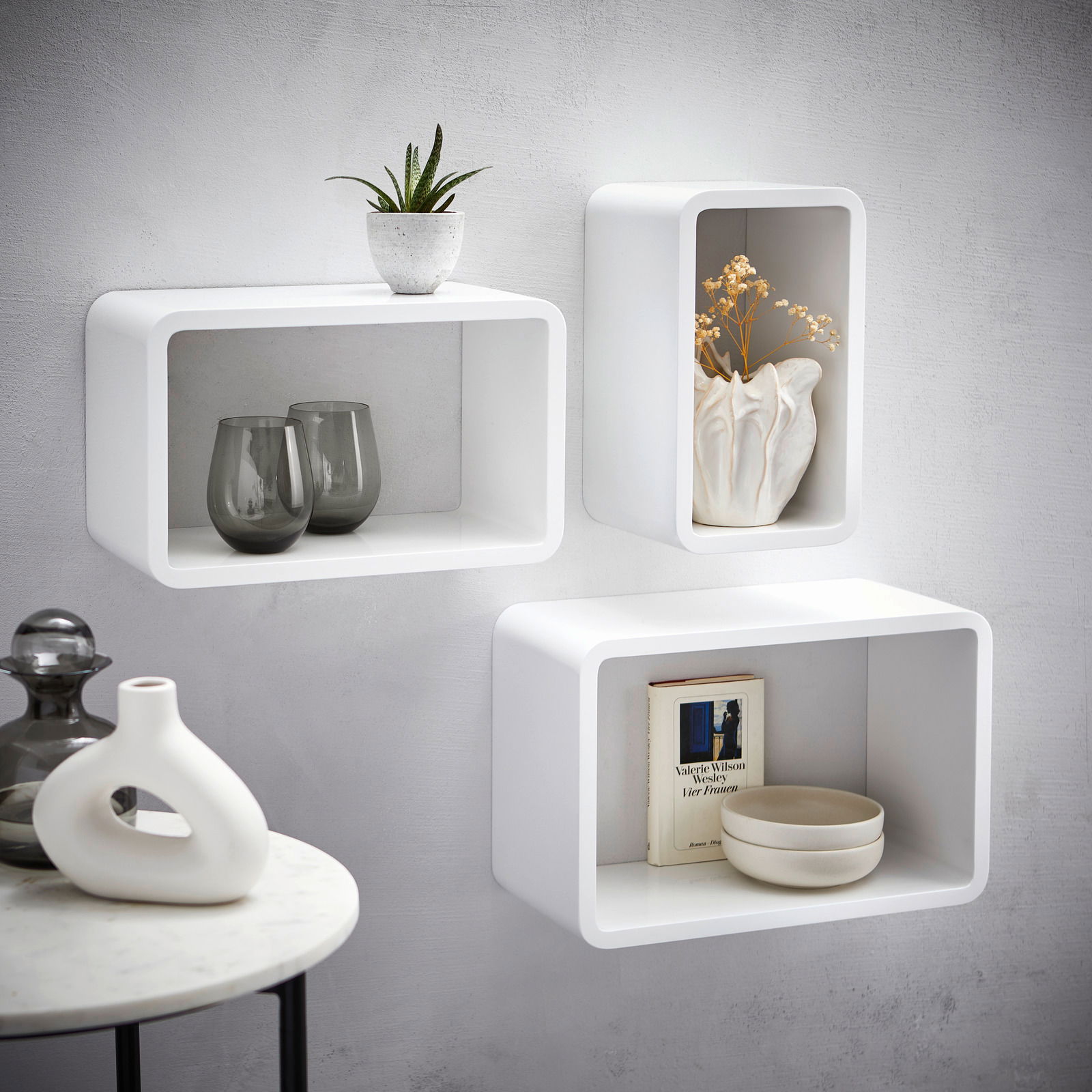 Wandregal-3er-Set in Weiß, bestehend aus drei rechteckigen Regalen, dekoriert mit Pflanzen, Vasen und Büchern, an einer grauen Wand montiert, seitliche Perspektive.
