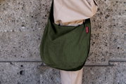 Olivgrüne, gerippte Schultertasche aus Stoff mit rotem Label, getragen über der Schulter an einer Person in beiger Kleidung vor einer Steinwand, seitliche Nahaufnahme.
