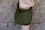 Olivgrüne, gerippte Schultertasche aus Stoff mit rotem Label, getragen über der Schulter an einer Person in beiger Kleidung vor einer Steinwand, seitliche Nahaufnahme.