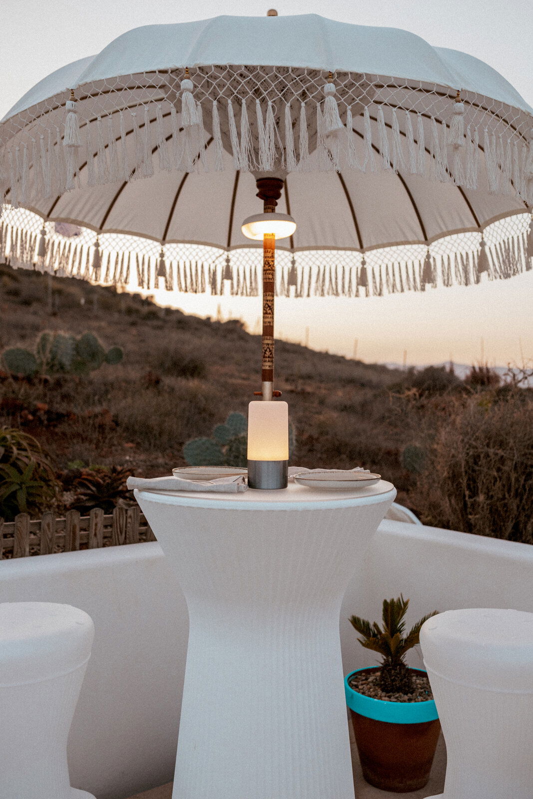 Großer weißer Sonnenschirm mit Fransen und Makramee-Details über einem runden Stehtisch mit warm leuchtender Lampe, aufgenommen in leichter Untersicht von vorn auf einer Terrasse bei Sonnenuntergang.