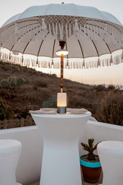 Großer weißer Sonnenschirm mit Fransen und Makramee-Details über einem runden Stehtisch mit warm leuchtender Lampe, aufgenommen in leichter Untersicht von vorn auf einer Terrasse bei Sonnenuntergang.