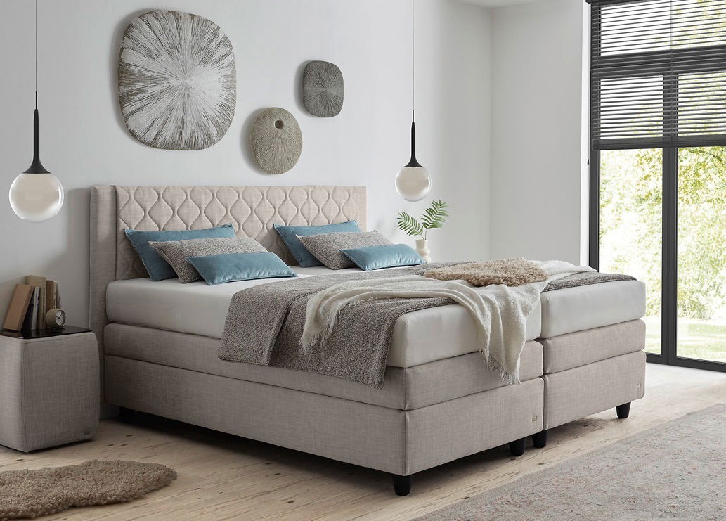 Boxspringbett in einem modernen Schlafzimmer, seitliche Perspektive, mit beigen Polstern, blauen Kissen und dekorativen Wandobjekten.