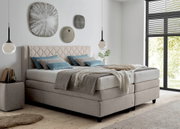 Boxspringbed in een moderne slaapkamer, gezien vanaf de zijkant, met beige bekleding, blauwe kussens en decoratieve wandobjecten.