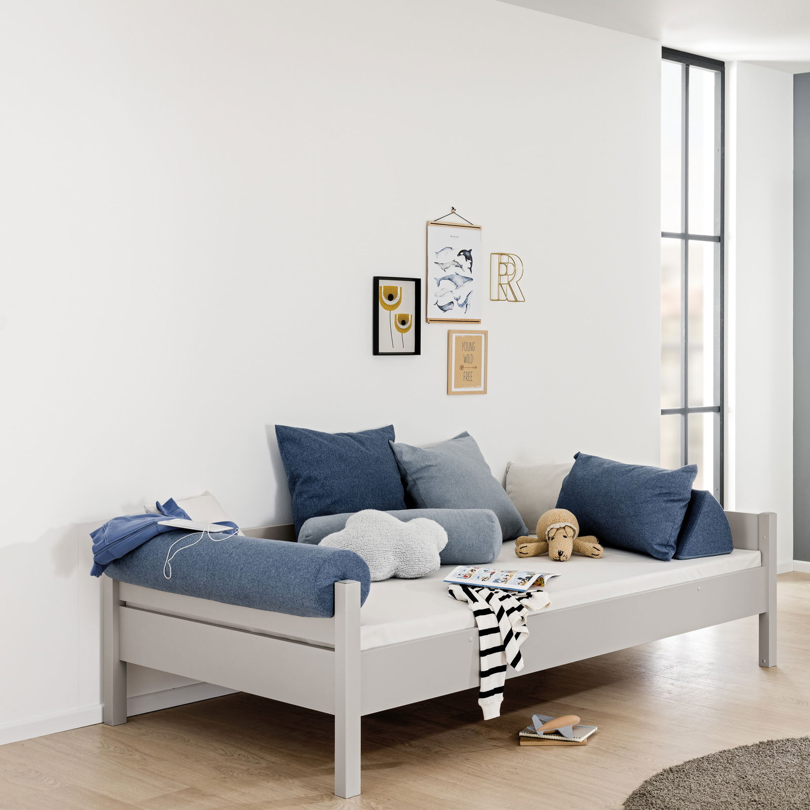 Modernes graues Bett mit blauen und grauen Kissen, seitliche Perspektive, dekoriert mit Wandbildern und Spielzeug.