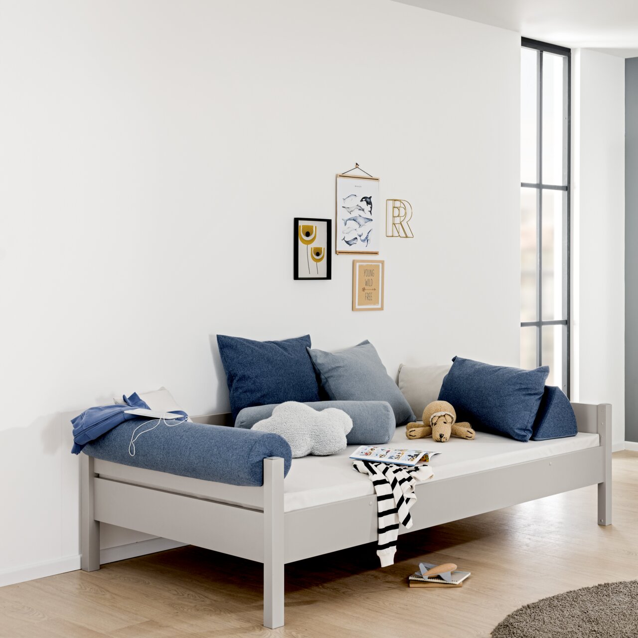 Modernes graues Bett mit blauen und grauen Kissen, seitliche Perspektive, dekoriert mit Wandbildern und Spielzeug.