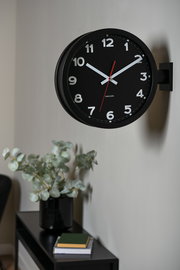 Schwarze, doppelseitige Wanduhr mit weißen Ziffern und rotem Sekundenzeiger, seitlich an der Wand montiert.