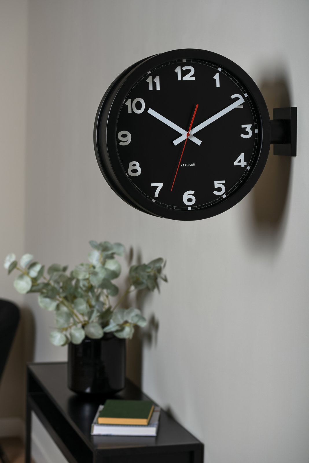 Schwarze, doppelseitige Wanduhr mit weißen Ziffern und rotem Sekundenzeiger, seitlich an der Wand montiert.