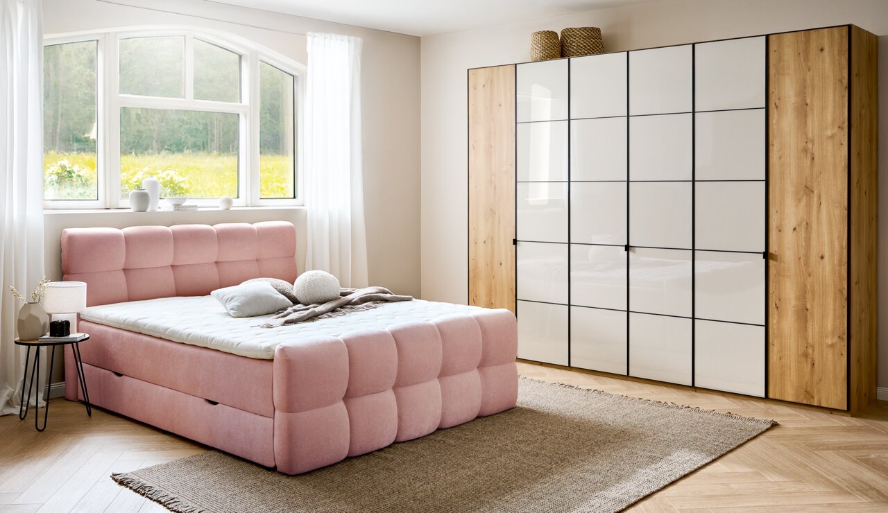 Rosa Boxbett in einem hellen Schlafzimmer mit großem Fenster, Holzboden und modernem Kleiderschrank, seitliche Perspektive.