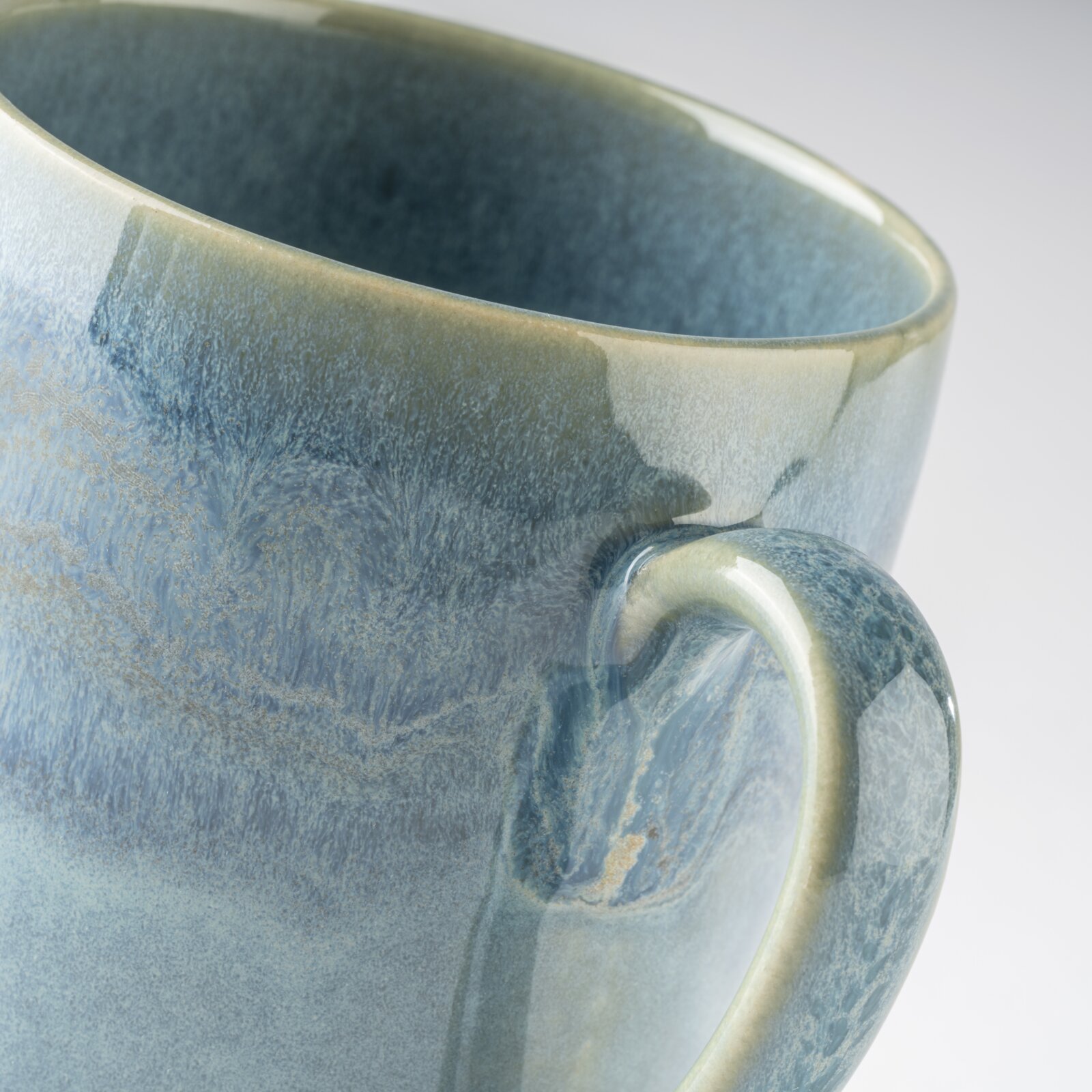 Detailaufnahme einer blau-grau glasierten Keramiktasse mit Henkel, schräg von oben aus seitlicher Perspektive vor hellem Hintergrund