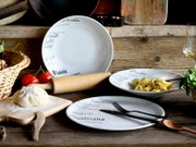 Rustikales Stillleben mit weißem, italienisch beschriftetem Porzellan-Geschirrset (tiefer Teller/Schüssel und flache Teller) auf Holztisch, dazu Tortellini, Besteck, Teig, Nudelholz und Tomaten; Perspektive: leicht von oben, frontal schräg.