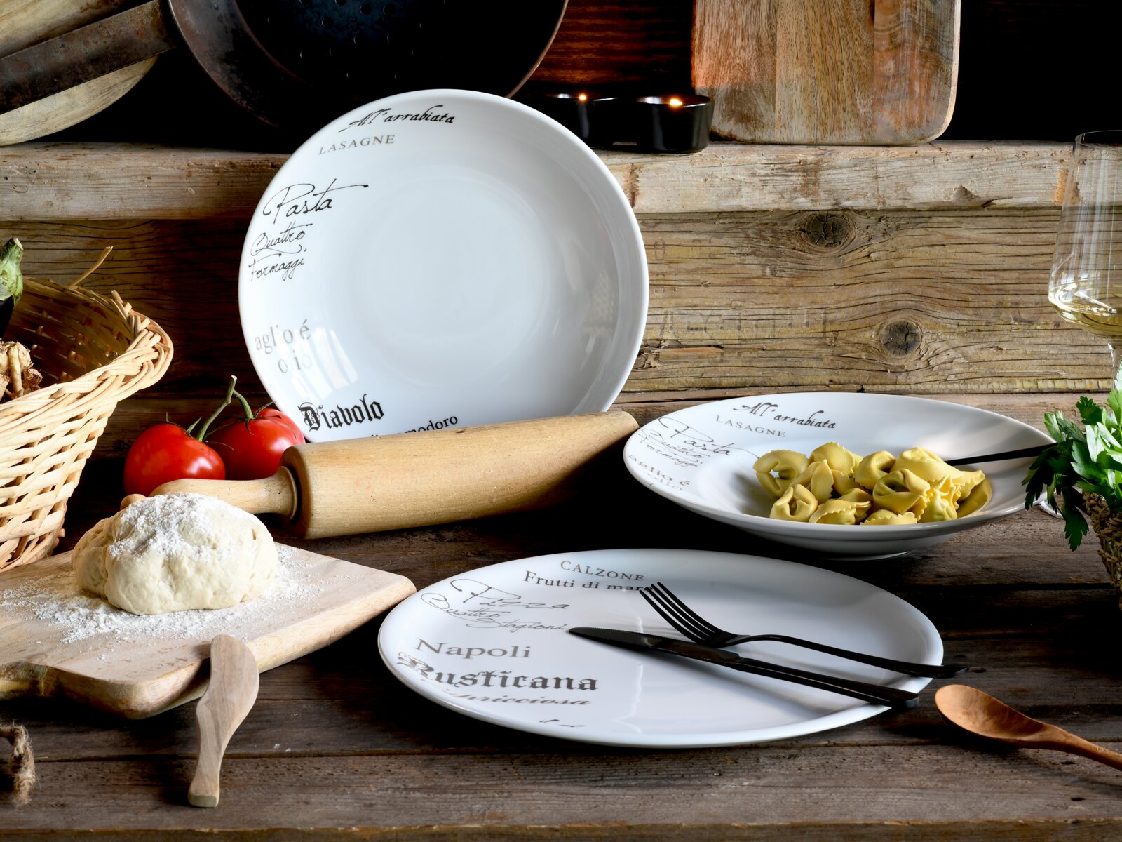 Rustikales Stillleben mit weißem, italienisch beschriftetem Porzellan-Geschirrset (tiefer Teller/Schüssel und flache Teller) auf Holztisch, dazu Tortellini, Besteck, Teig, Nudelholz und Tomaten; Perspektive: leicht von oben, frontal schräg.