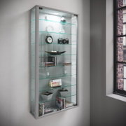 Wandmontierte Glasvitrine mit silbernem Rahmen, Glasböden und LED-Beleuchtung, bestückt mit Deko und Büchern; Ansicht schräg von vorne links.