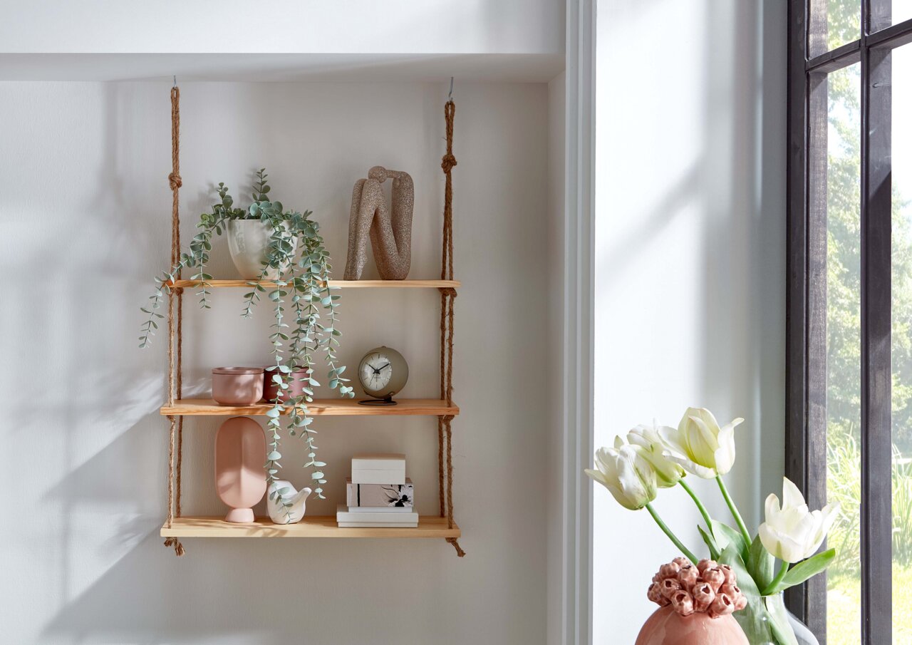 Wandhängeregal aus hellem Holz mit drei Ablagen an Seilen, dekoriert mit Pflanze, Vasen, Uhr und Boxen; seitliche Perspektive im hellen Raum neben Fenster und Tulpenvase.