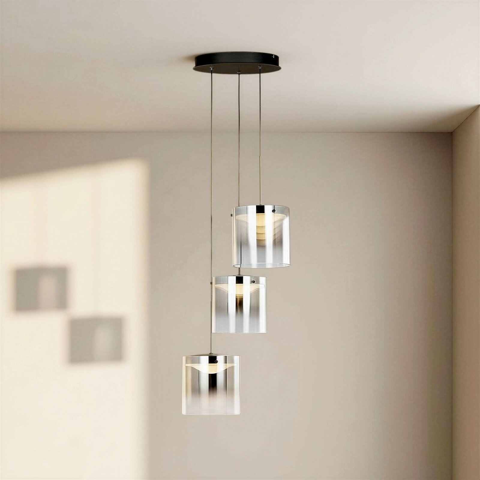 Moderne hanglamp met drie lampen, voorzien van drie cilindervormige, transparante glazen kappen en een zwarte plafondplaat, opgehangen in een minimalistische ruimte, vooraanzicht licht van onderaf