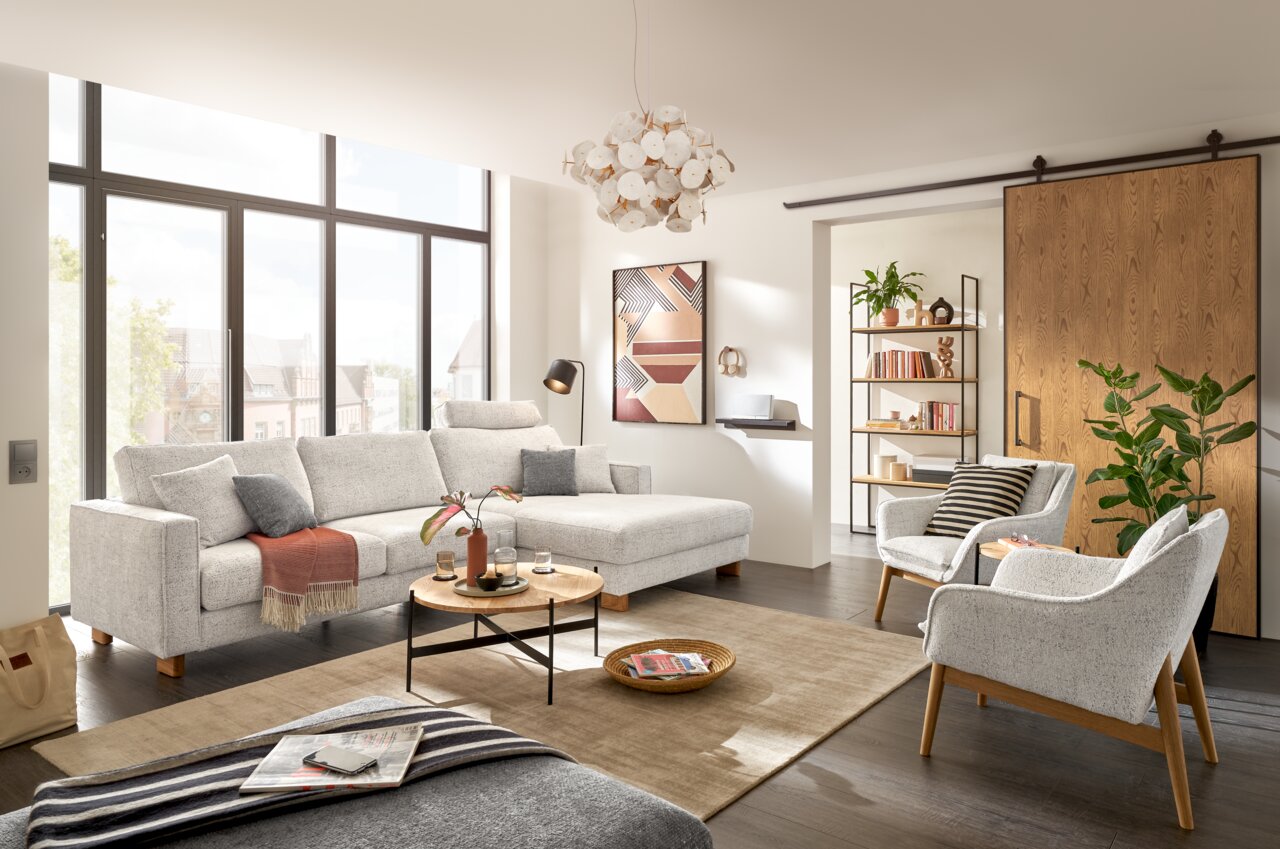 Moderner Wohnraum mit einem hellgrauen Sessel im Vordergrund, umgeben von einem großen Fenster, einem Sofa und einem Couchtisch. Perspektive von vorne.