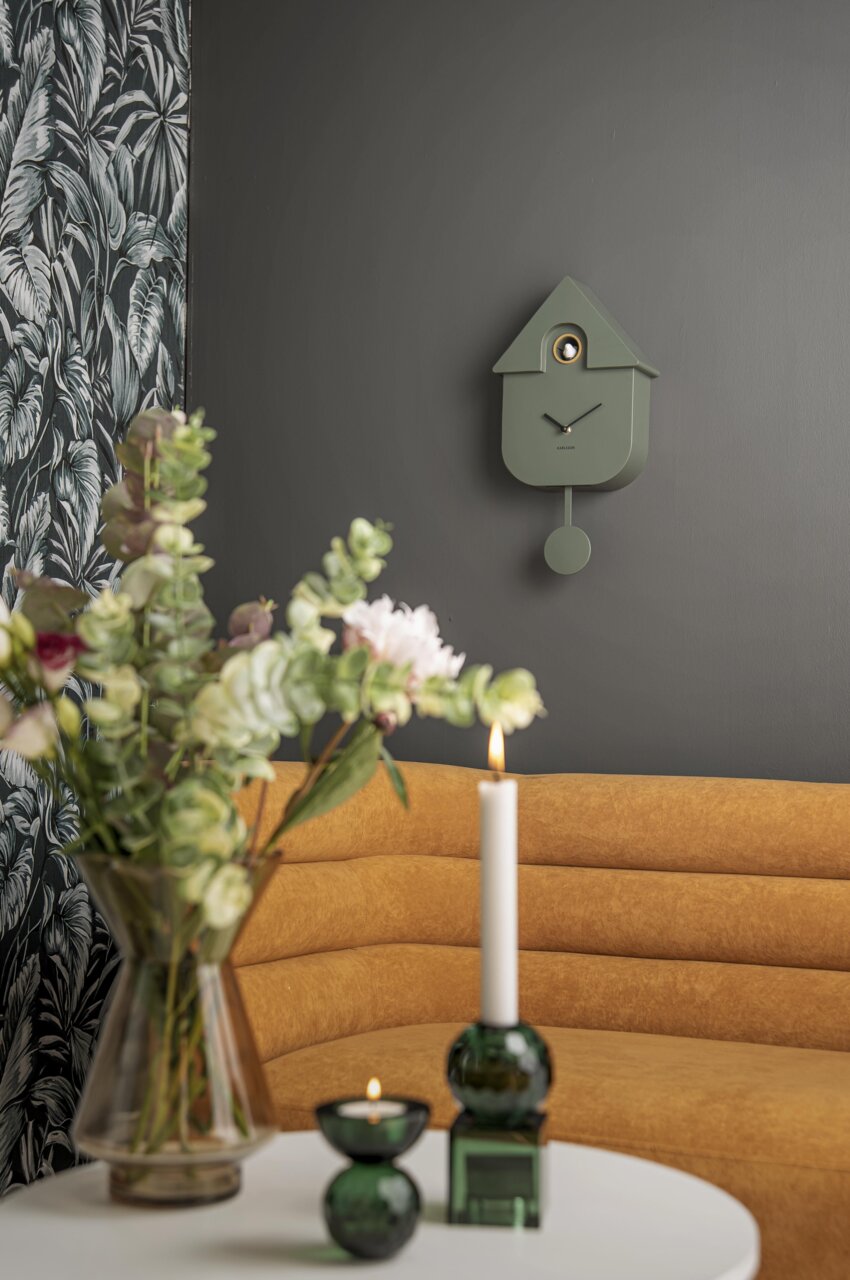 Moderne grüne Wanduhr in Kuckucksuhren-Design an einer dunkelgrauen Wand, mit einem gelben Sofa und dekorativen Elementen im Vordergrund, seitliche Perspektive.