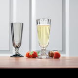 Zwei elegante Sektkelche aus der Opera-Serie, einer gefüllt mit Sekt, auf einem Tisch mit Erdbeeren und einem Korken, aus seitlicher Perspektive fotografiert.