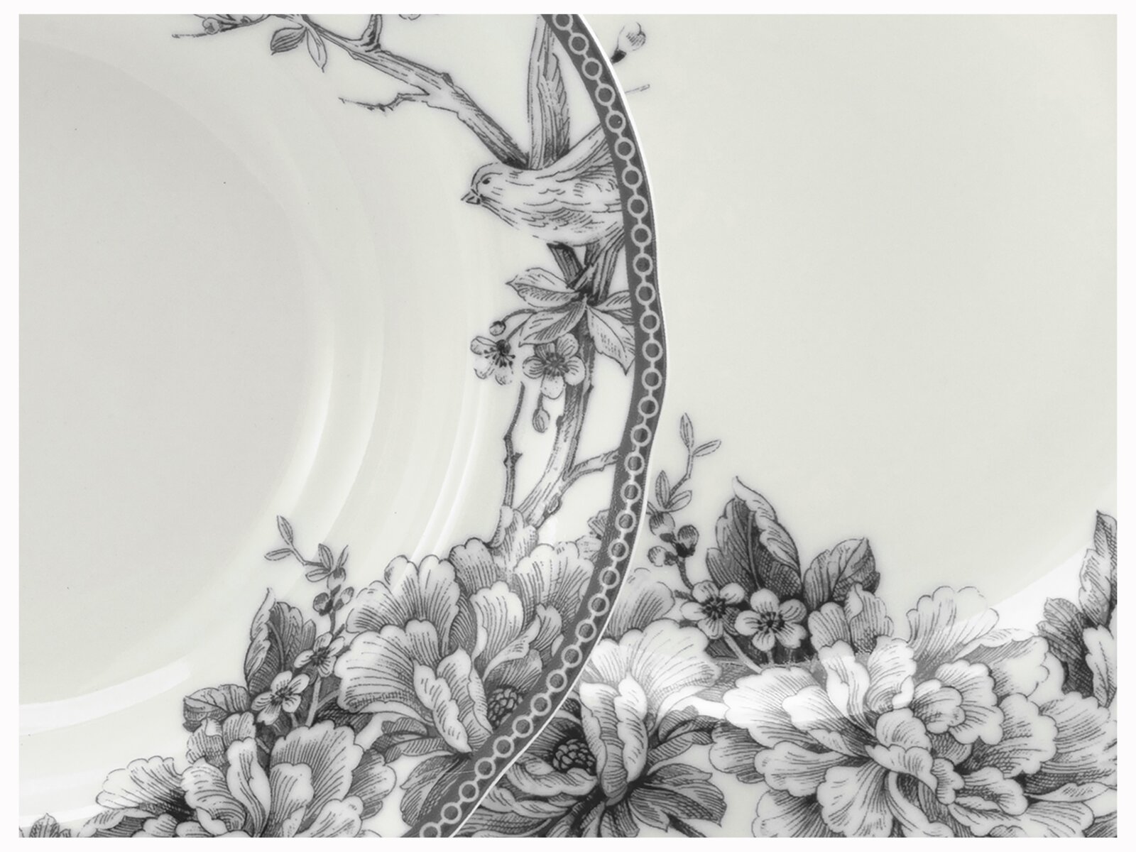 Detailaufnahme aus leicht schräger Draufsicht: weißer Porzellanteller mit schwarzem floralen Dekor, Vogelmotiv am Rand und ornamentalem Bordürenmuster.
