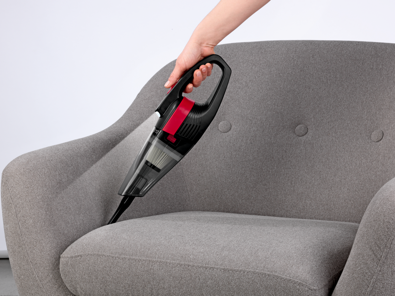 Akku Handstaubsauger in der Hand gehalten, reinigt einen grauen Sessel aus seitlicher Perspektive.