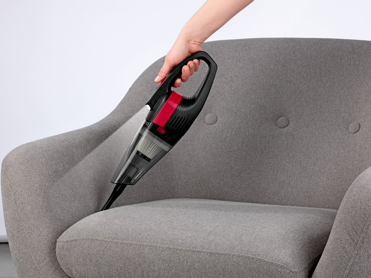 Akku Handstaubsauger in der Hand gehalten, reinigt einen grauen Sessel aus seitlicher Perspektive.