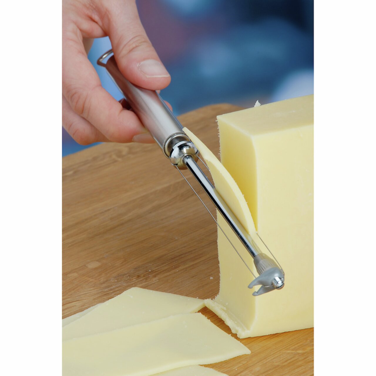 Nahaufnahme eines Käseschneiders PROFI PLUS, der eine dünne Scheibe von einem Käseblock schneidet. Der Käseschneider hat einen ergonomischen Griff aus Edelstahl.