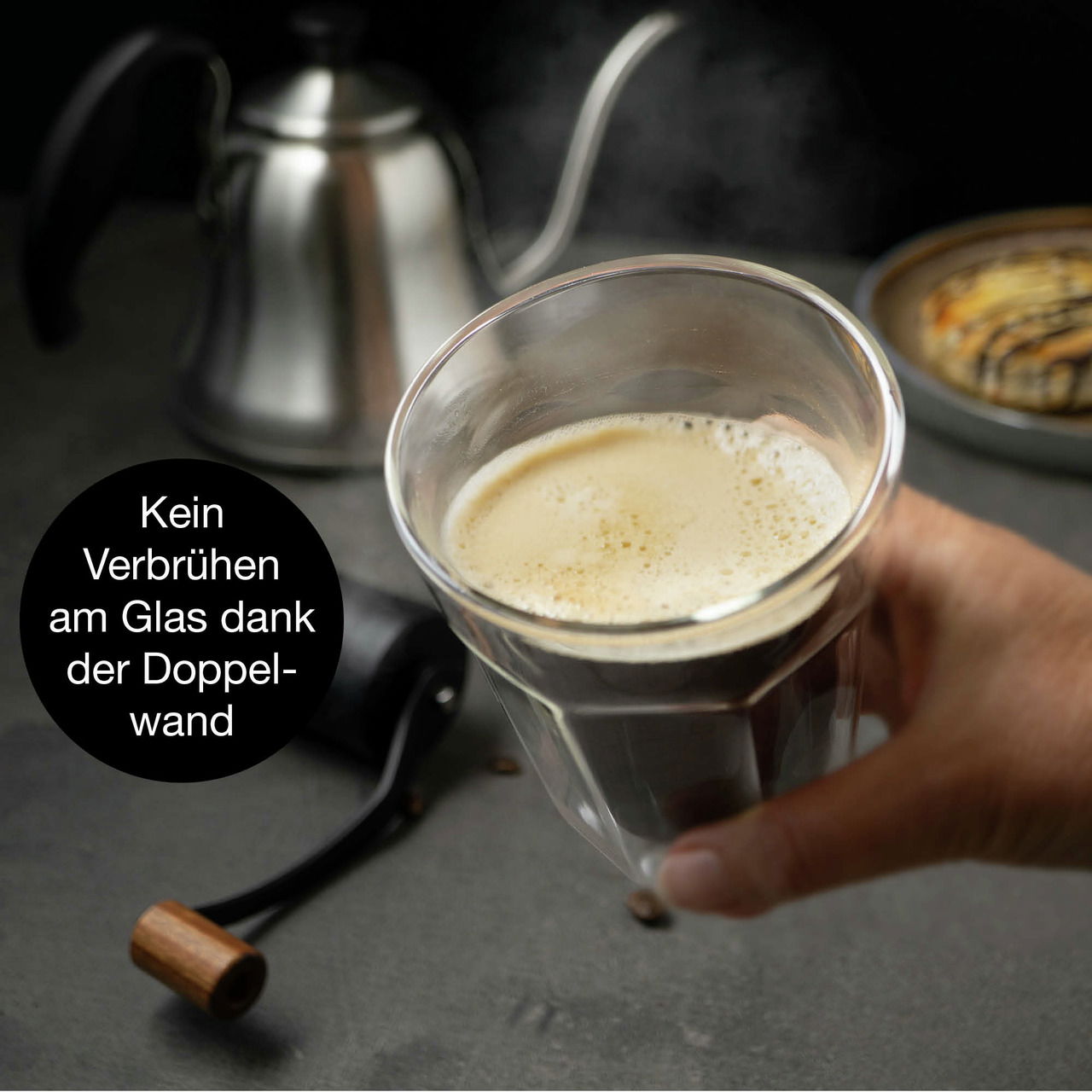 Hand hält ein doppelwandiges Glas mit Kaffee, im Hintergrund eine Kaffeekanne und ein Gebäck, seitliche Perspektive.