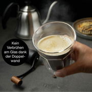 Hand hält ein doppelwandiges Glas mit Kaffee, im Hintergrund eine Kaffeekanne und ein Gebäck, seitliche Perspektive.