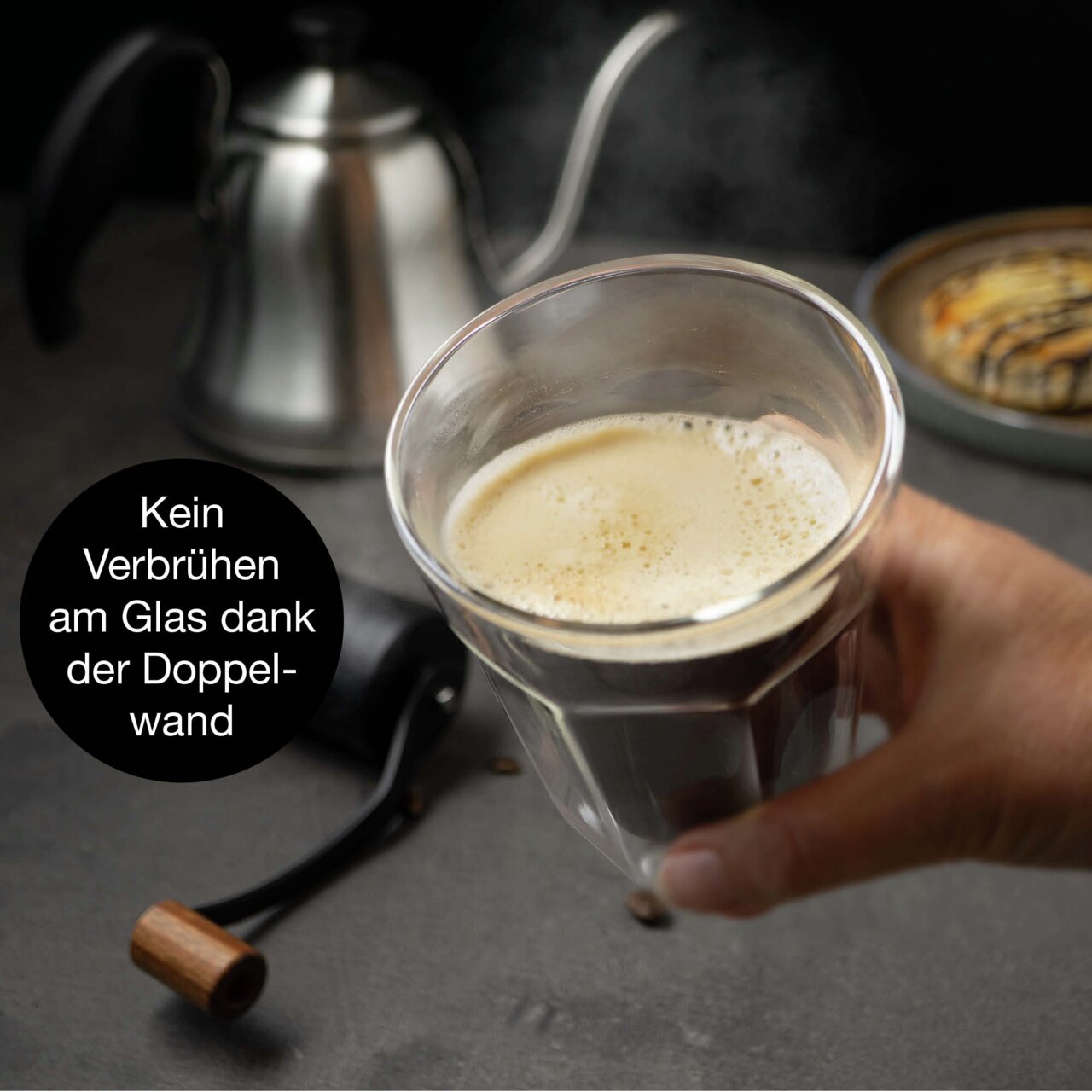 Een hand houdt een dubbelwandig glas met koffie vast; op de achtergrond staan een koffiepot en een gebakje; zijaanzicht.