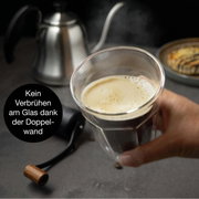 Een hand houdt een dubbelwandig glas met koffie vast; op de achtergrond staan een koffiepot en een gebakje; zijaanzicht.