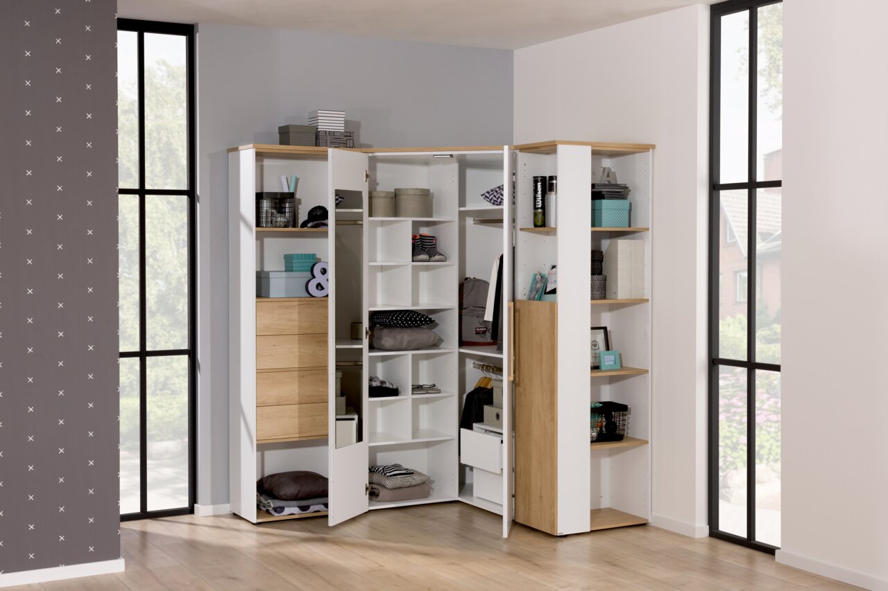 Eck-Kleiderschrank in heller Holzoptik mit offenen und geschlossenen Fächern, gefüllt mit verschiedenen Gegenständen, aus einer frontalen Perspektive fotografiert.