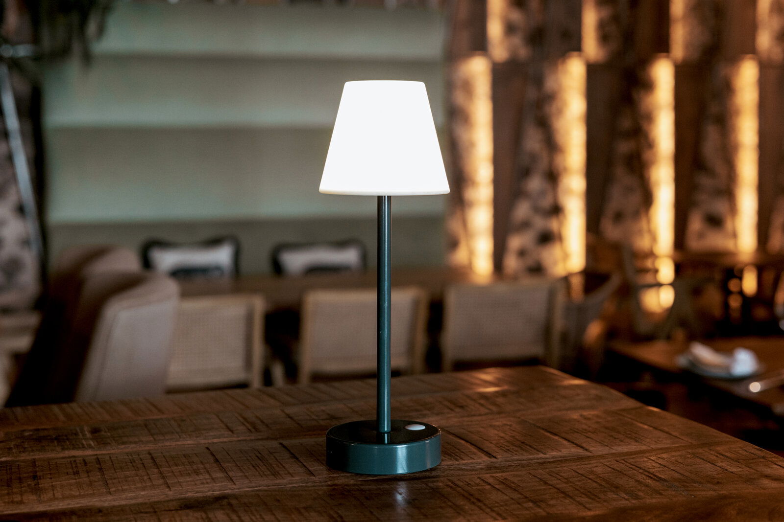 Moderne kabellose Tischlampe mit weißem Lampenschirm und schwarzem Standfuß auf einem Holztisch in einem Restaurant, frontal aus Augenhöhe fotografiert.