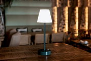 Moderne kabellose Tischlampe mit weißem Lampenschirm und schwarzem Standfuß auf einem Holztisch in einem Restaurant, frontal aus Augenhöhe fotografiert.