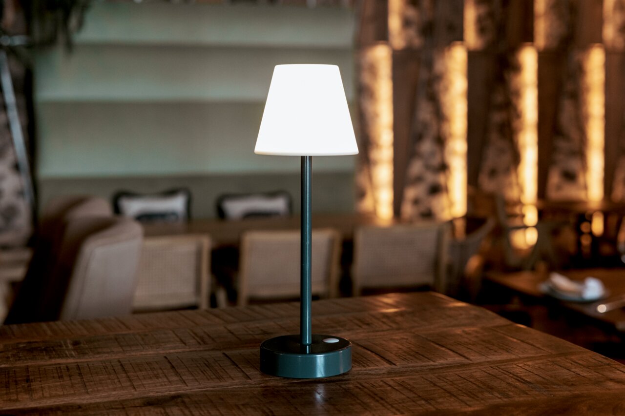 Moderne kabellose Tischlampe mit weißem Lampenschirm und schwarzem Standfuß auf einem Holztisch in einem Restaurant, frontal aus Augenhöhe fotografiert.