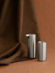 Moderner Seifenspender aus Edelstahl, seitliche Perspektive, vor braunem Stoffhintergrund