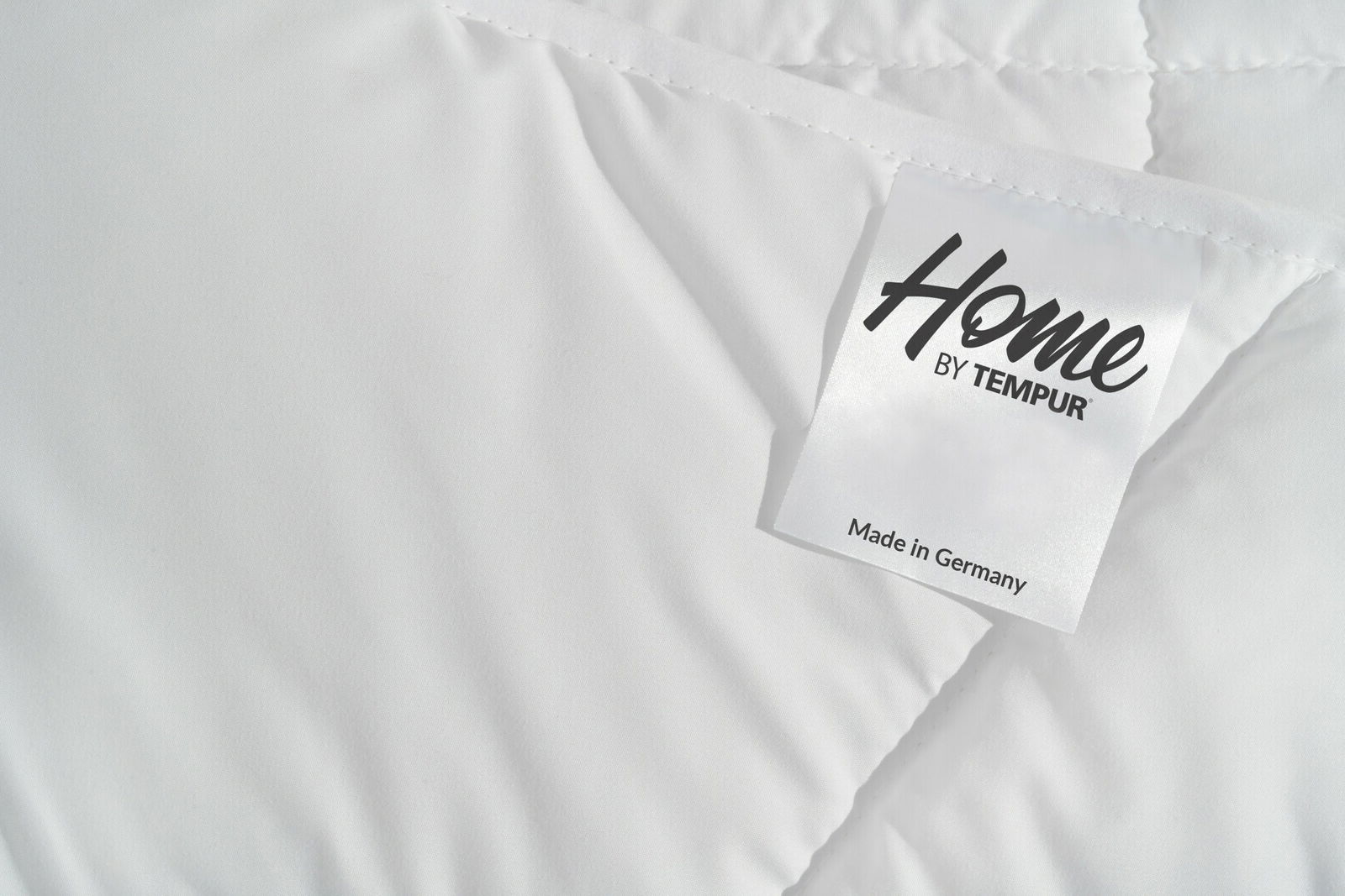 Nahaufnahme einer weißen, gesteppten Decke mit einem Etikett, das 'Home by Tempur' zeigt, perspektivisch von oben.