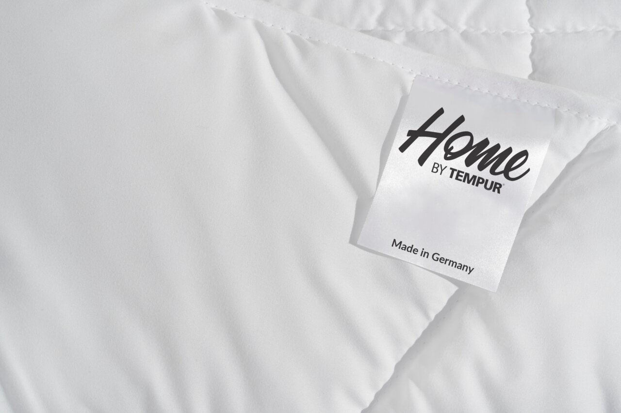 Nahaufnahme einer weißen, gesteppten Decke mit einem Etikett, das 'Home by Tempur' zeigt, perspektivisch von oben.