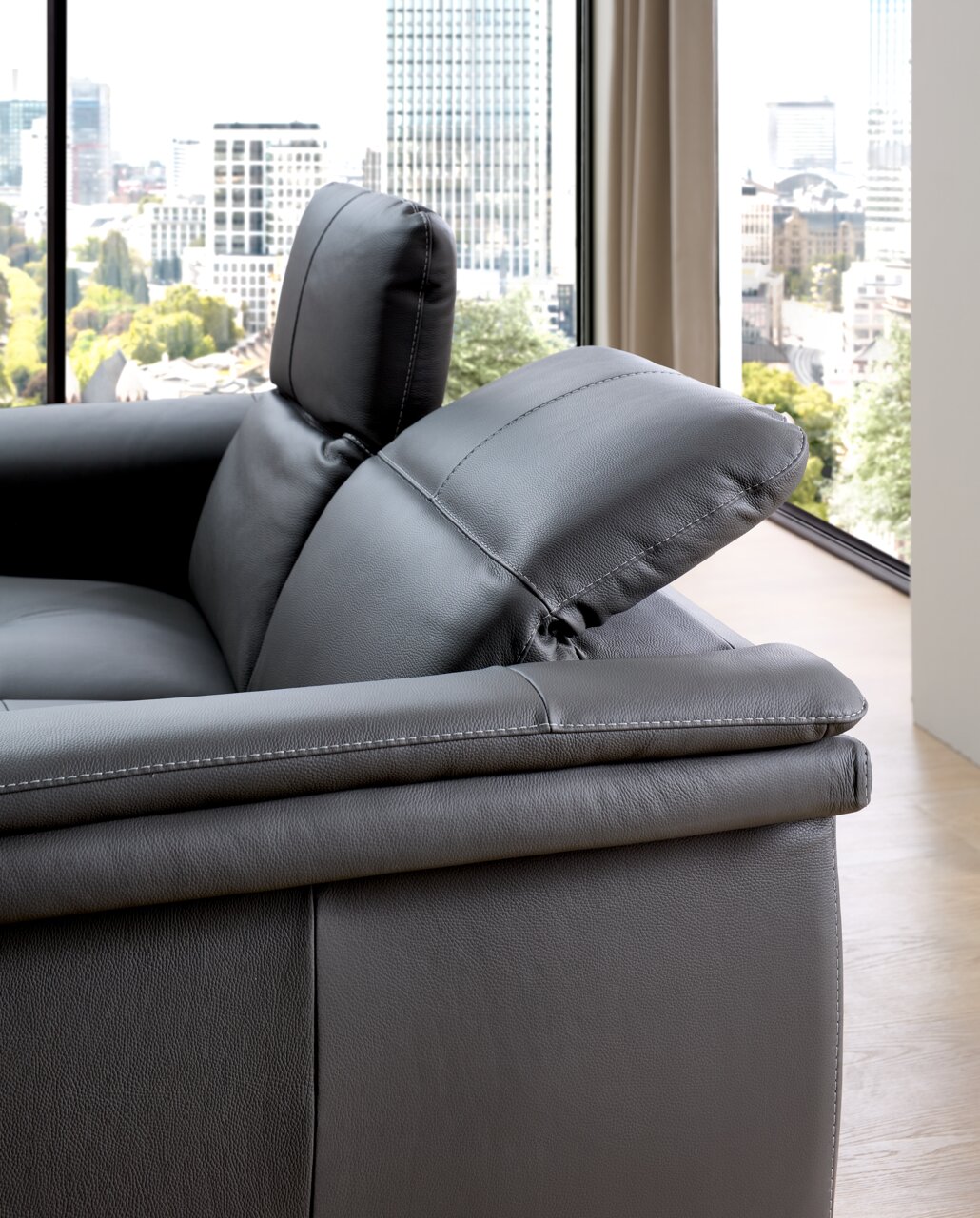 Nahaufnahme eines grauen Ledersofas mit verstellbarer Kopfstütze, seitliche Perspektive mit Blick auf eine Stadtlandschaft durch ein großes Fenster.