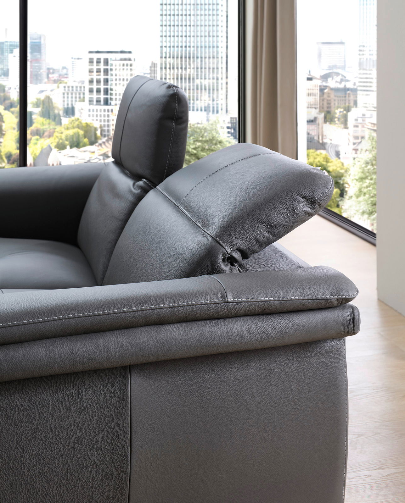 Nahaufnahme eines grauen Ledersofas mit verstellbarer Kopfstütze, seitliche Perspektive mit Blick auf eine Stadtlandschaft durch ein großes Fenster.