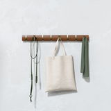 Wandgarderobe aus Holz mit mehreren Haken, an denen ein grünes Seil, eine beige Tasche und ein grünes Tuch hängen. Frontalansicht.