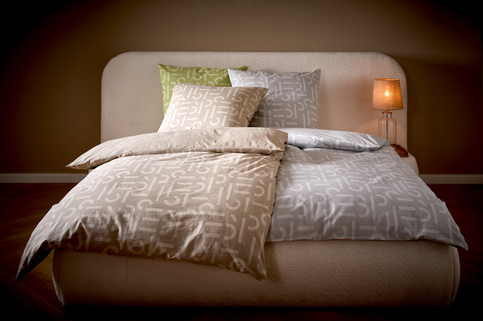Modernes Bett mit zweifarbiger Bettwäsche in Beige und Grau mit grafischem Muster, mehreren Kissen und leuchtender Nachttischlampe rechts, Frontalansicht aus leicht erhöhter Perspektive