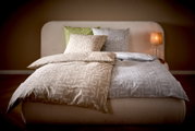 Modernes Bett mit zweifarbiger Bettwäsche in Beige und Grau mit grafischem Muster, mehreren Kissen und leuchtender Nachttischlampe rechts, Frontalansicht aus leicht erhöhter Perspektive