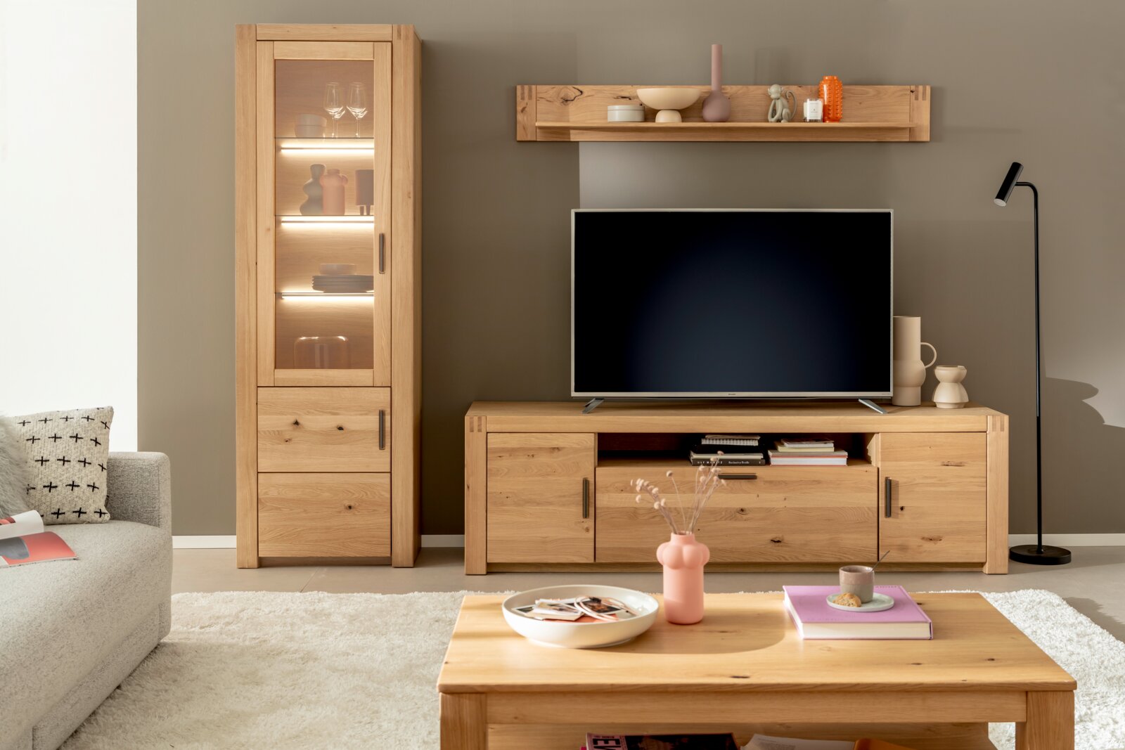 Wohnzimmeransicht mit einem modernen TV-Element aus Holz, bestehend aus einem TV-Schrank, einem hohen Vitrinenschrank und einem Wandregal. Der TV-Schrank hat offene Fächer und Schubladen, der Vitrinenschrank ist beleuchtet und zeigt dekorative Objekte. Pe