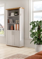 Hohes Regal mit weißer Holzoptik und naturfarbener Abdeckplatte, offenen Fächern und zwei Türen unten, dekoriert mit Büchern und Ordnern, im hellen Raum am Fenster; Perspektive: leicht schräg von vorn links