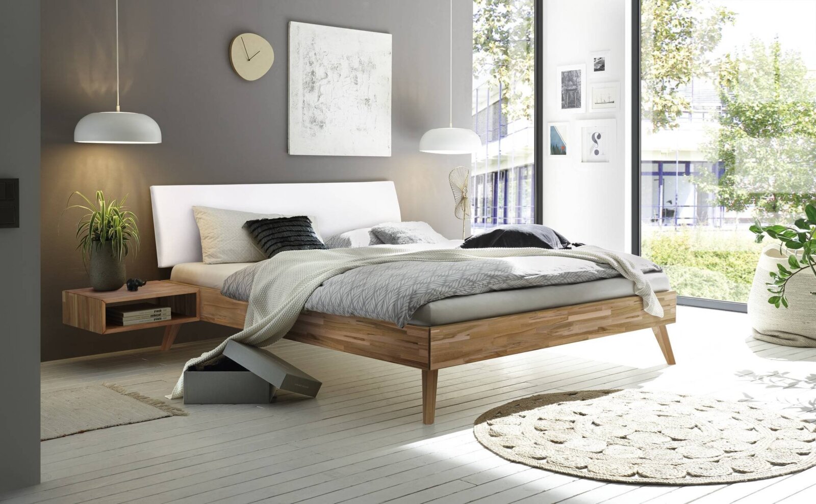 Modern houten bedframe met wit hoofdeinde, gezien vanaf de zijkant, omringd door stijlvolle decoratie in een lichte kamer met grote ramen.