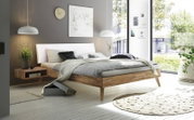 Modern houten bedframe met wit hoofdeinde, gezien vanaf de zijkant, omringd door stijlvolle decoratie in een lichte kamer met grote ramen.