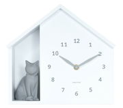 Weiße Wanduhr in Form eines Hauses mit einer grauen Katzenfigur auf der linken Seite, frontal betrachtet.