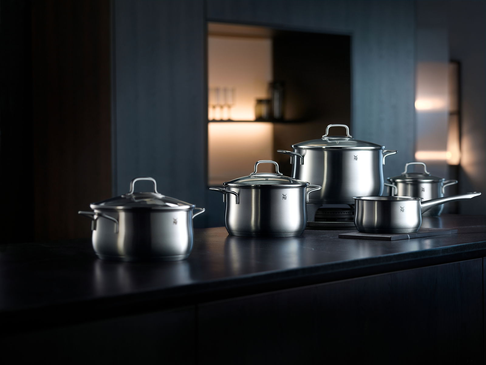 Topfset Brillant 4-tlg. aus Edelstahl, bestehend aus drei Töpfen und einer Stielkasserolle, auf einer dunklen Küchenarbeitsplatte in moderner Umgebung, seitliche Perspektive.