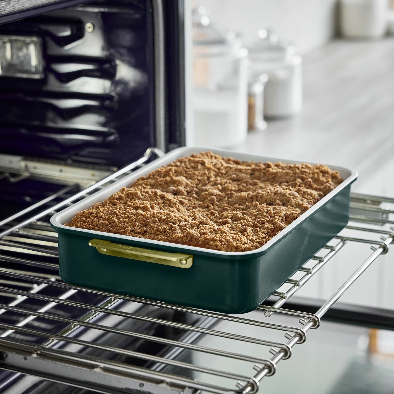 Rechteckige Backform mit goldenen Griffen, gefüllt mit einem gebackenen Kuchen, in einem geöffneten Ofen aus seitlicher Perspektive.